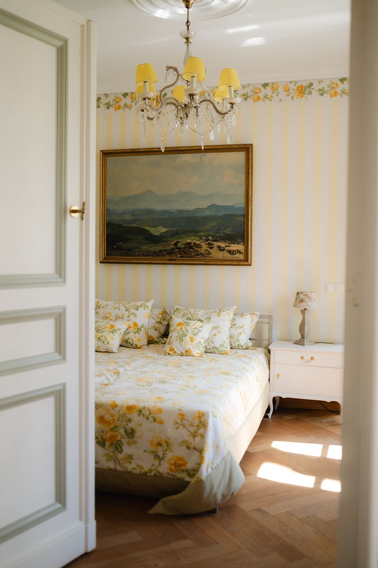 Blick durch eine geöffnete weiße Tür in ein gelb gestreiftes Schlafzimmer mit Kristalllüster