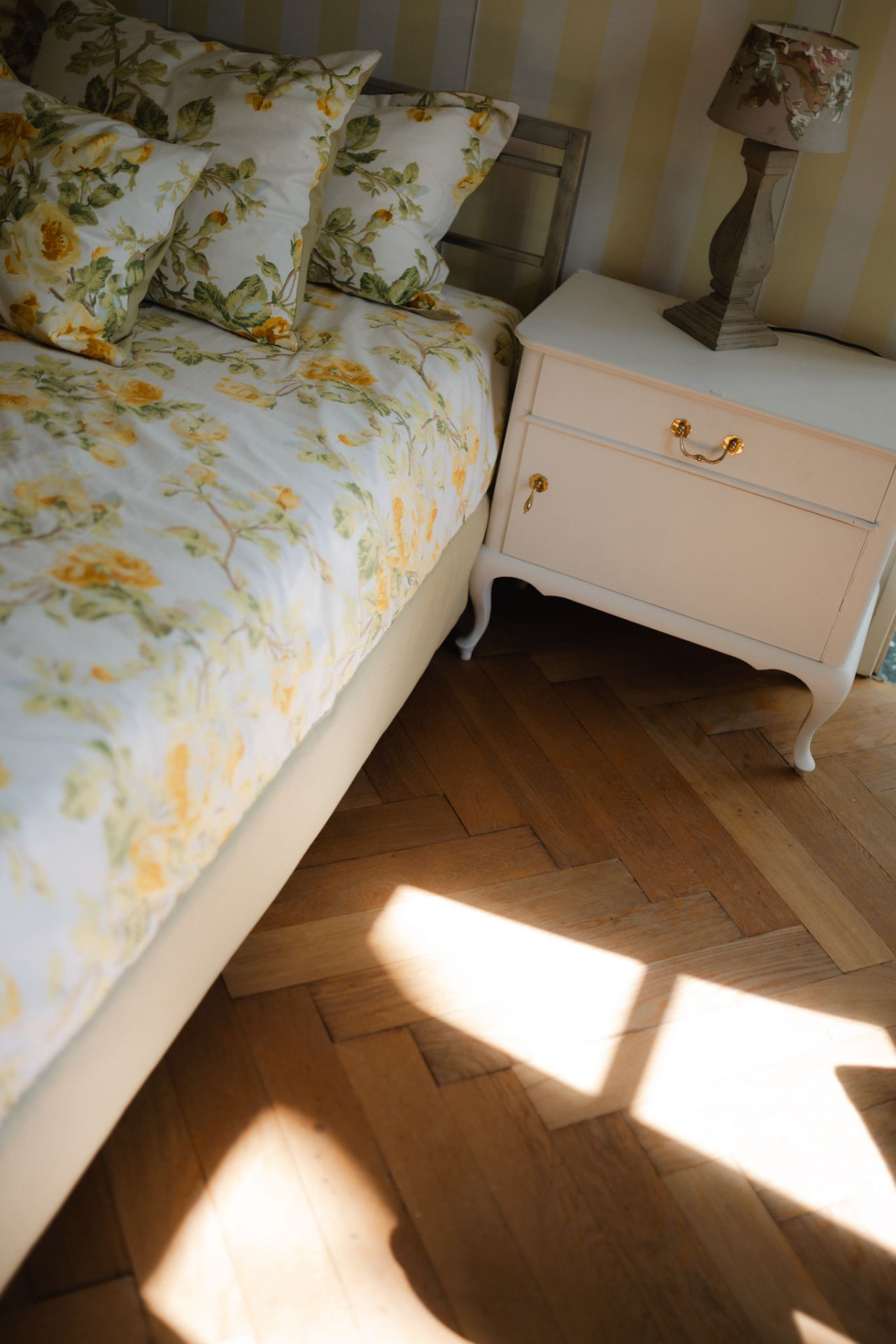Bett mit gelb geblümter Bettwäsche neben einem weißen Nachttisch mit Lampe, gelb gestreifte Tapete im Hintergrund