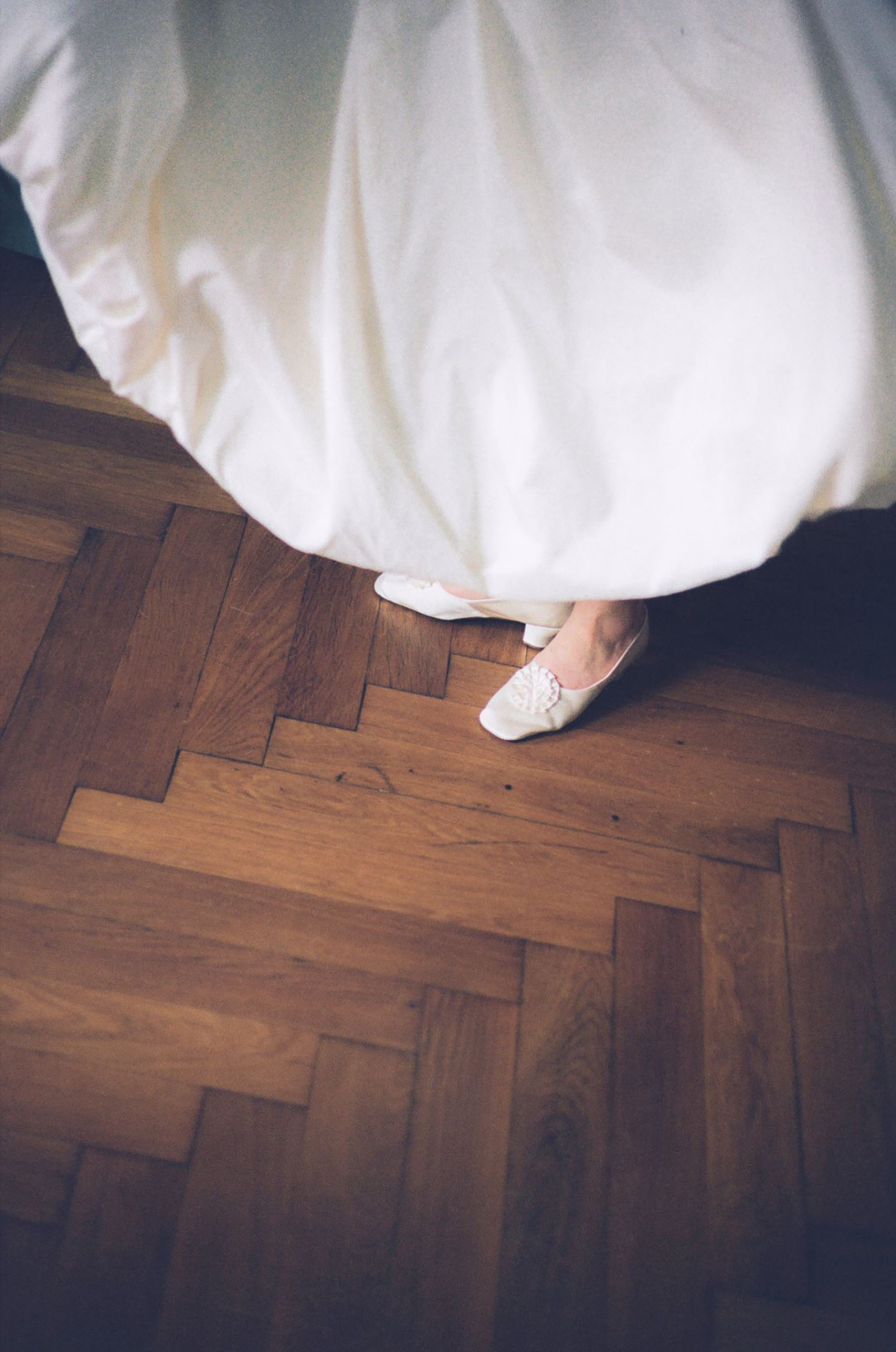 Brautschuhe mit Stoffrosette unter dem Saum eines weißen Hochzeitskleides, aufgenommen von oben auf einem Parkettboden