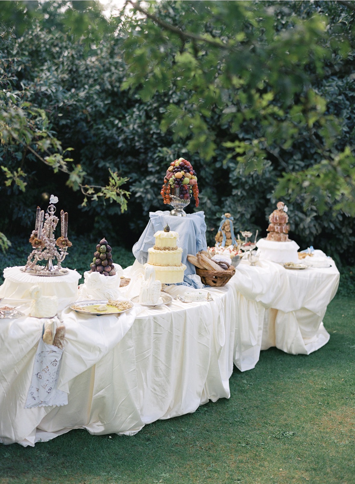 Desserttafel in weißer Draperie mit Torte unter Fruchtkrone, Croquembouche und Gebäck vor dunkelgrünen Hecken