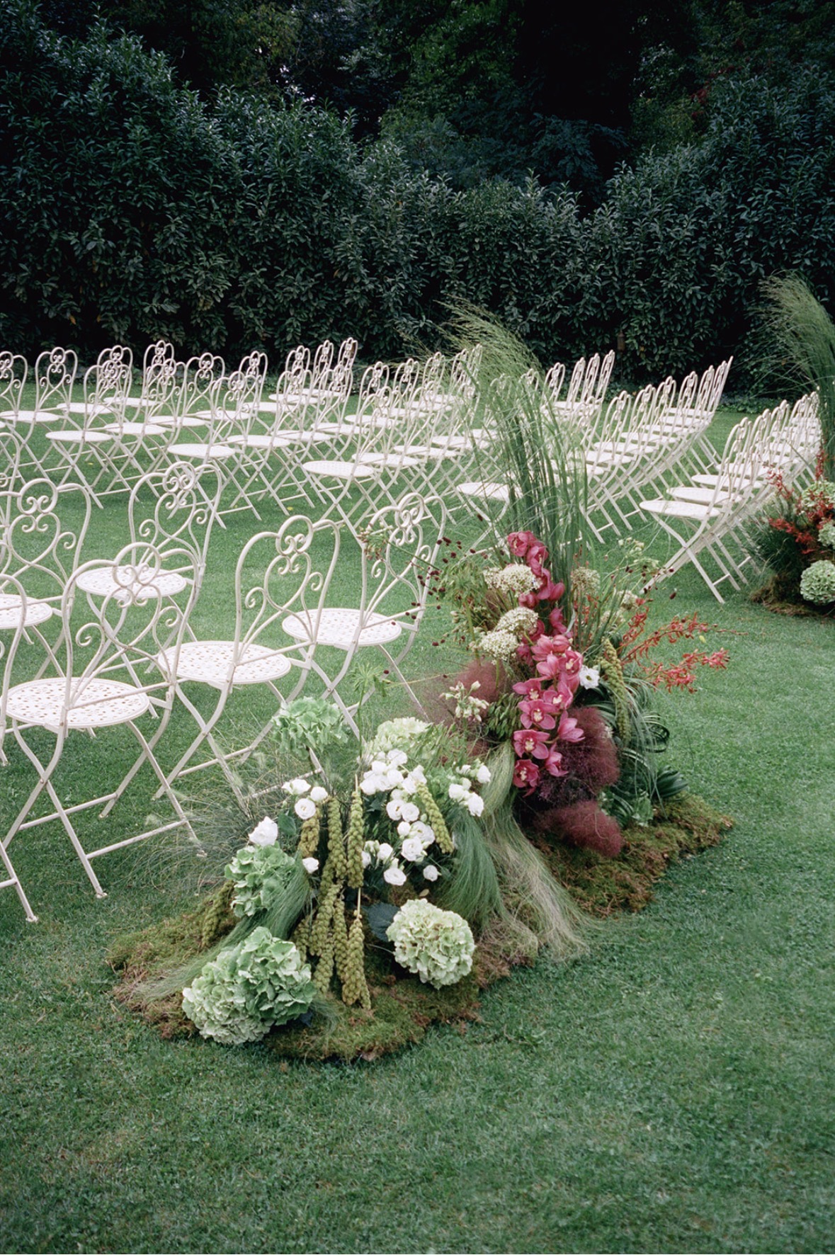 Wilde Blumenarrangements mit rosa Orchideen, Gräsern und Moos zwischen Reihen weißer Klappstühle