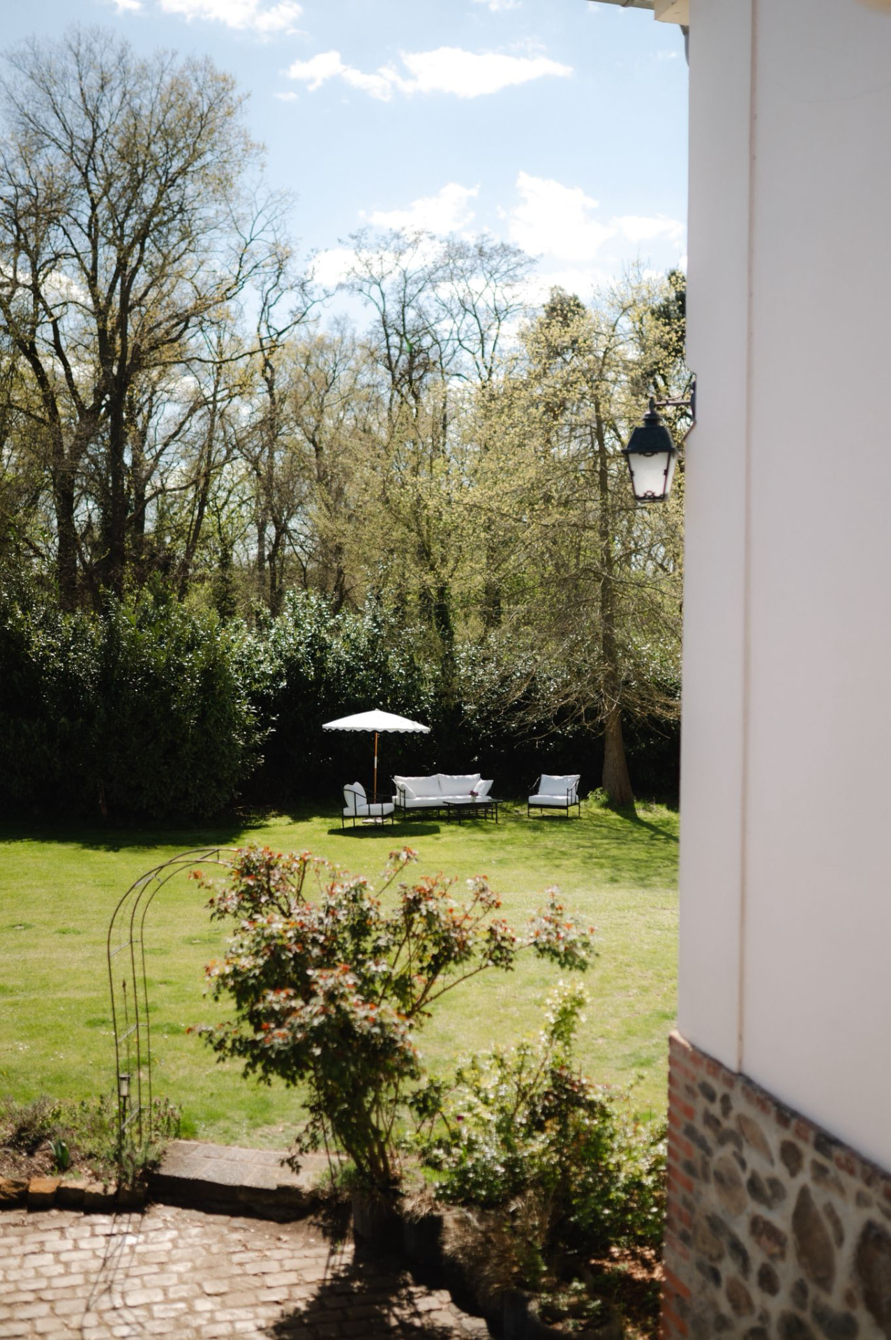 Weißes Sofa mit Kissen unter weißem Sonnenschirm auf dem Rasen, im Vordergrund ein Rosenbogen
