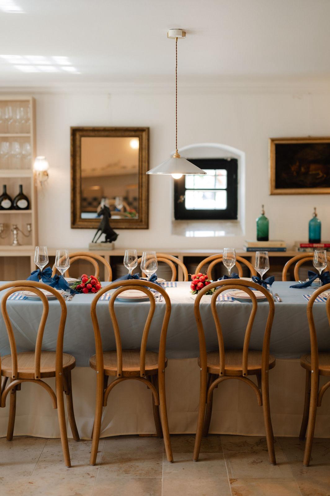 Gedeckter Tisch mit hellblauer Tischdecke, Holzstühlen und Weingläsern mit dunkelblauen Bändern, dahinter Weinregale und ein goldener Spiegel