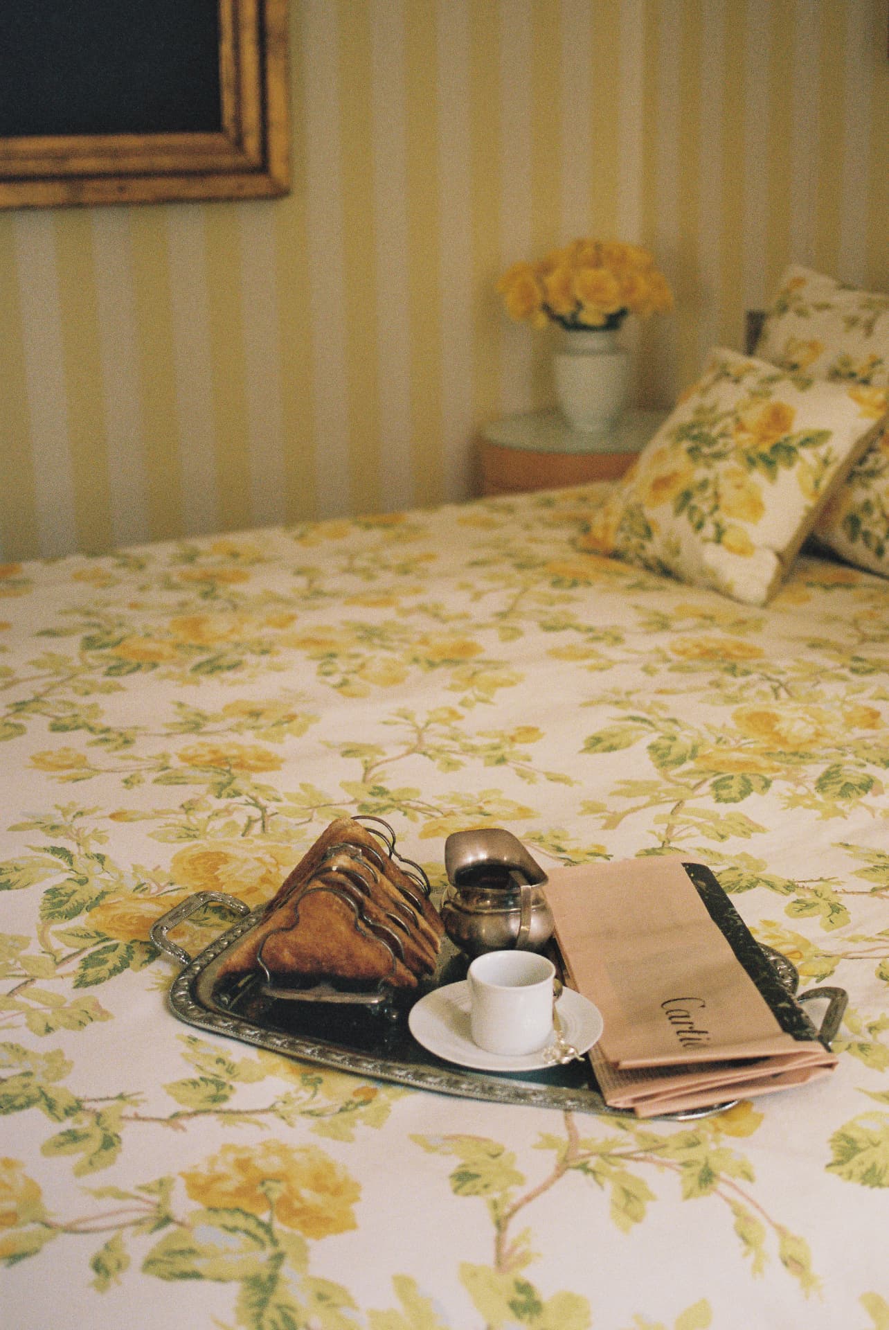 Versilbertes Tablett mit Toast, Kännchen und Zeitung auf einem Bett mit gelb geblümter Tagesdecke